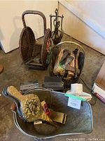Photo showing woven firewood baskets, fireplace tools, brass shovel, two small heaters and a white small fan grouped together on garage floor.
