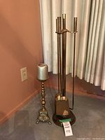 Wide view showing brass fireplace tool set with four tools and ornate candle holder with silver candle positioned near a corner wall.