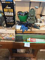 Top view of gardening table with garden tools (hose sprayers, pruners), small water fountain statue, green bucket with gloves and hand tools, boxed gutter guard, and boxed solar lantern, plus garden supply packages in drawer.