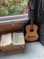 Valencia acoustic guitar standing next to three boxes of assorted sheet music placed on the floor near a window.