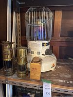 Full view of the kerosene heater and two small brass lanterns on a shelf with a cardboard box.