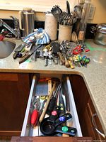 Overview of assorted kitchen utensils and tools spread across kitchen sink and countertop plus kitchen drawer with more utensils and measuring cups.
