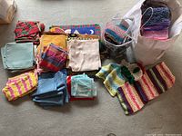 Photo showing assorted table linens including napkins and placemats in multiple colors and patterns, plus knit scarves in bags and laid out.