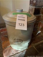 Photo of the vintage Medalta stoneware crock with lid against brick background, showing label with lot number and the Medalta stamp visible on the crock body.