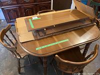 Dining table with four chairs arranged around it, wooden furniture in medium brown finish with two table leaves on top, taped measurement label on table.