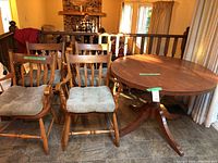 Photo showing four wooden chairs with vertical slat backs and beige seat cushions next to a circular wooden table in used condition.
