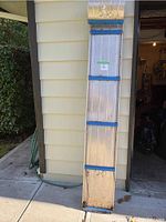 Full length view of metal toboggan propped vertically against house wall, showing the overall condition with surface wear, rust, blue support braces, and rope handles.
