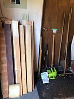 Photo showing assorted wood pieces of various lengths and thicknesses stacked vertically next to door, and several garden tools including sledgehammer, axe, shovel, broom, and Poulan electric chainsaw on floor.