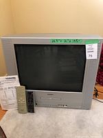 Front view of the Toshiba 20-inch CRT combination television with DVD/CD player, two remotes and user manual placed in front. Green tape on top with measurement details.