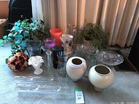 Overall assortment of vases and artificial plants on carpet in front of closed window curtains.