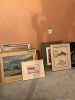 Six framed artworks leaning against wall on carpet, shows variety of frame styles and sizes.