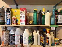 Wide view of garage shelf with multiple garden and yard spray containers of various sizes and brands, showing condition and contents