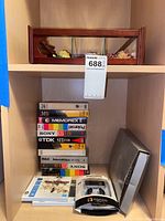 Wooden shelving with wooden display case of ceramic roses on top shelf. Stack of 9 VHS tapes including Memorex, Sony, TDK, RCA, Polaroid. Unopened SanDisk Sansa slot music player in retail box with wired earbuds. Polaroid boxed item beside them.
