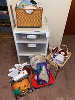 Photo showing white plastic three-drawer storage bin with clear front drawers and white handles, woven basket containing various crafting supplies on top, medium-sized globe on the floor, red wagon holding a boxed ceramic talking Jack-o'-lantern pumpkin and a ghost wooden figure nearby, and a separate basket holding a vintage rag doll.