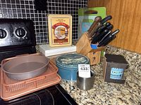 Overview of countertop with bakeware, tins, knife block, cutting boards on granite surface near stove.