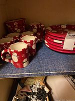 Side shelf with six red Christmas-themed plates stacked, six matching bowls, and six cups with snowmen and holiday motifs visible