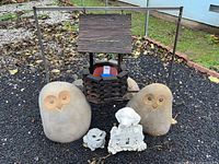Front view of the two large stone owl statues, wooden wishing well planter, small lamb statue, and white cherub statue grouped on black gravel.