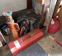 Photo shows orange hydraulic jack, red toolbox, jumper cables, gas can, and vehicle jack placed on a dusty floor against wooden wall