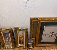 Photo of seven framed floral wall art paintings of various sizes with gold ornate frames leaning against a wall on a wooden floor.
