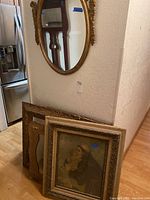 Photo showing three wall art frames leaning against a wall beneath the oval mirror. One contains a religious print of a saint and child, two other frames appear empty or with unclear images.