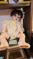 Front view of ceramic doll seated with dark curly hair, lace bonnet, cream dress, socks and shoes.