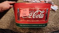 Front of Coca-Cola metal tin showing main logo and two bottle images on a red background with green bottom border.