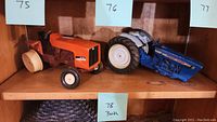 Two farm tractor models on shelf, tagged 75 (orange) and 76 (blue) side by side.