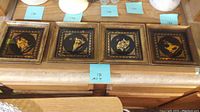 Four square decorative frames set in a row on a table, showing gold-tone zodiac signs on black backgrounds with ornate borders.