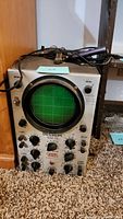 Front view of the oscilloscope showing circular green grid screen and multiple black control knobs. Unit has Tektronix branding below the screen.
