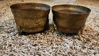 Two small brass pots side by side on carpet, showing aged patina and tripod feet.