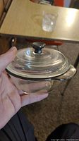 Photo of small clear glass ovenware dish held by hand showing overall shape, handles and lid knob.