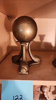 Solid brass stand with a spherical ball atop three fish-shaped legs, shown from front angle.