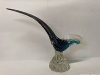Side profile showing the full body of the teal blue glass pheasant with long curved purple tail perched on a clear textured glass base.