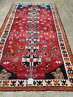 Full carpet laid out showing the red field, geometric motifs along central axis, and decorative orange, black, and white borders.