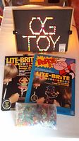Vintage Light Brite board lit with colored pegs to show the general shape and item condition, accompanied by two paper refill packs (Popeye and Bugs Bunny) and a bag of colored plastic light pegs.