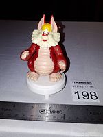 Front view of 1986 SNARF figure showing red and beige painted details, yellow tuft of hair, and facial expression. Mounted on white circular stand with ruler for scale showing figure height at about 3.5 inches.
