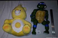 Front view of the yellow Care Bear plush and Teenage Mutant Ninja Turtle figure with ruler for scale.