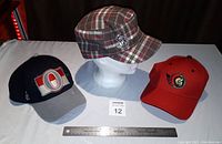 Photo of three Ottawa Senators hats: navy/grey baseball cap with large 'O' logo, red baseball cap with team logo, and plaid cadet style cap with metal emblem pin on side.