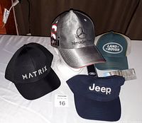 Four hats displayed on white surface: Matrix black cap, Mercedes-Benz silver cap with red and white '63', Land Rover green and gray mesh cap, Jeep navy visor.