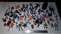 Overview photo showing about 49 mixed action figures and mini figurines laid out on a white surface with a ruler at bottom for scale.