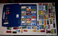 Overhead photo showing a large blue plastic carrying case partially filled with multiple rows of small vintage diecast cars and trucks in various colors, plus additional loose vehicles placed around the case.