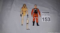 Two Kenner Star Wars action figures on white textured background. The 1977 Stormtrooper has detached legs positioned in front. The 1978 X Wing Pilot stands fully assembled.