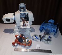 Image of the Star Wars vehicle lot showing the white AT-AT walker with one figure inside, the blue TIE Fighter, and two speeders with pilot figures, along with a ruler for scale.