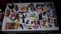 Overall view of all toys and collectibles on a white tablecloth, including multiple action figures, toy cars, and plush items.