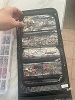Photo showing a hand holding open a black fabric holder displaying 6 clear zipper compartments with assorted small metal charms in various shapes and colors inside.