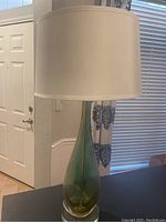 Side view of the glass table lamp showing the green teardrop glass base and white drum shade, placed near a window and door.