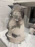 Front view of the cat pottery sculpture, showing the stylized face, incised lines for features, and natural black and tan smoke fired finish.