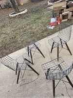 Four modern style chairs with wire mesh seats and backs, wooden legs, and black metal supports, shown outdoors on concrete surface.
