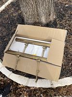 Two gold picture light fixtures packed in cardboard box on ground outdoors. One fixture is unpacked showing adjustable arms; the other remains partially boxed.