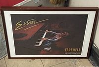 Framed Elton John Farewell Yellow Brick Road tour poster leaning against a wall outdoors, showing the entire piece and the frame's condition.